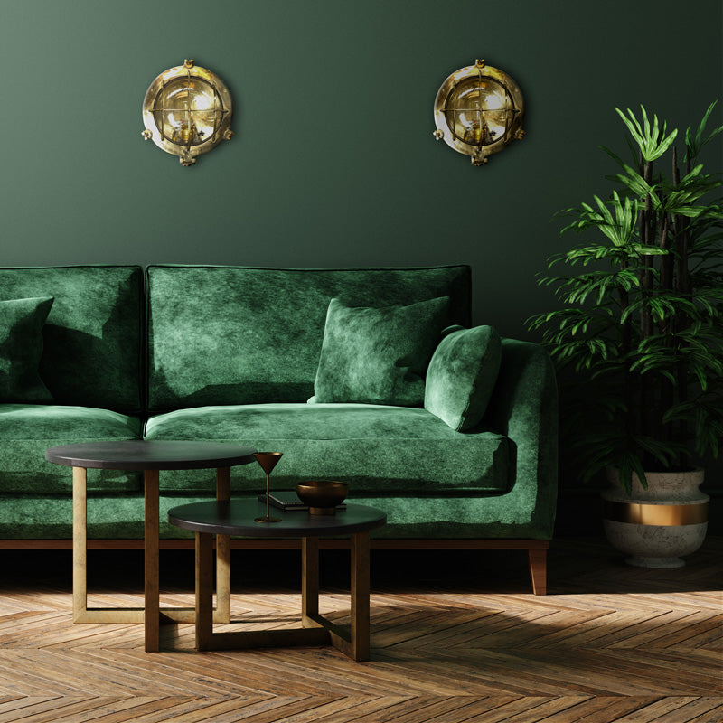 Vintage industrial brass bulkhead wall lighting refurbished, mounted on a living room wall with dark green wall, green velvet sofa and large green plant. Modern industrial style living room