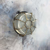 A vintage industrial brass circular bulkhead wall light with target style protective cage over a prismatic glass shade.