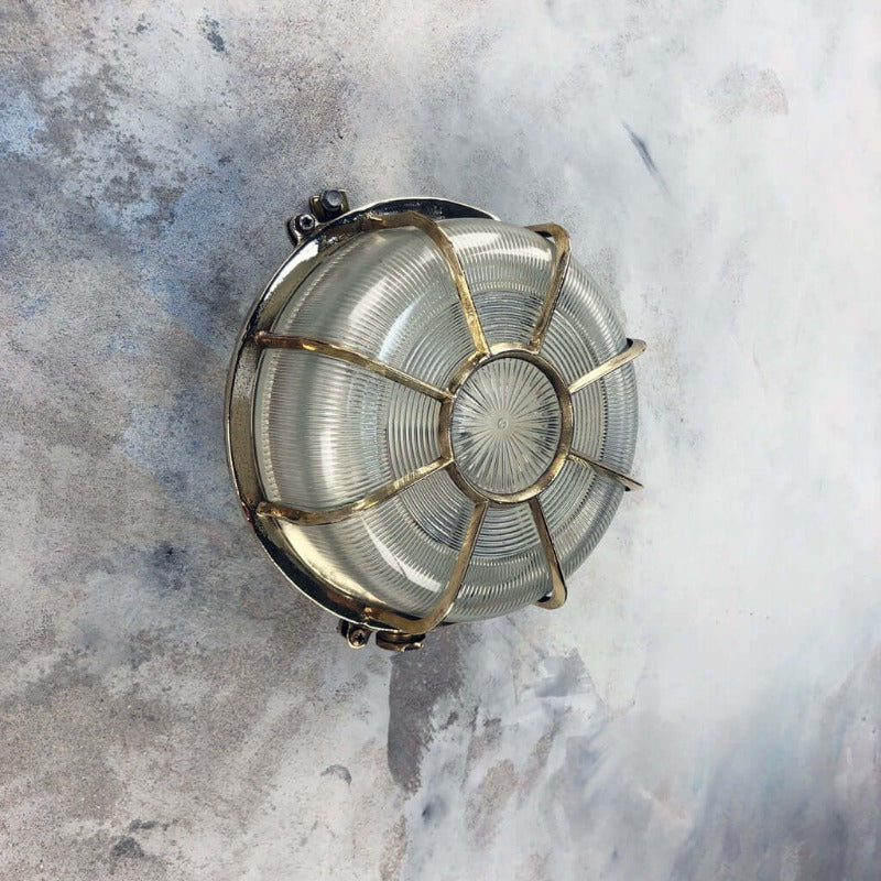 A vintage industrial brass circular bulkhead wall light with target style protective cage over a prismatic glass shade. 